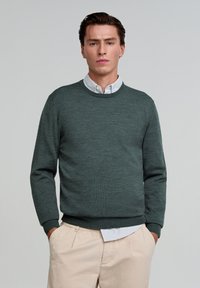 Donkergroen crewneck trui gemaakt van gestructureerde stof, met lange mouwen en een ribbelrand, gedragen over een lichtblauw overhemd met kraag.