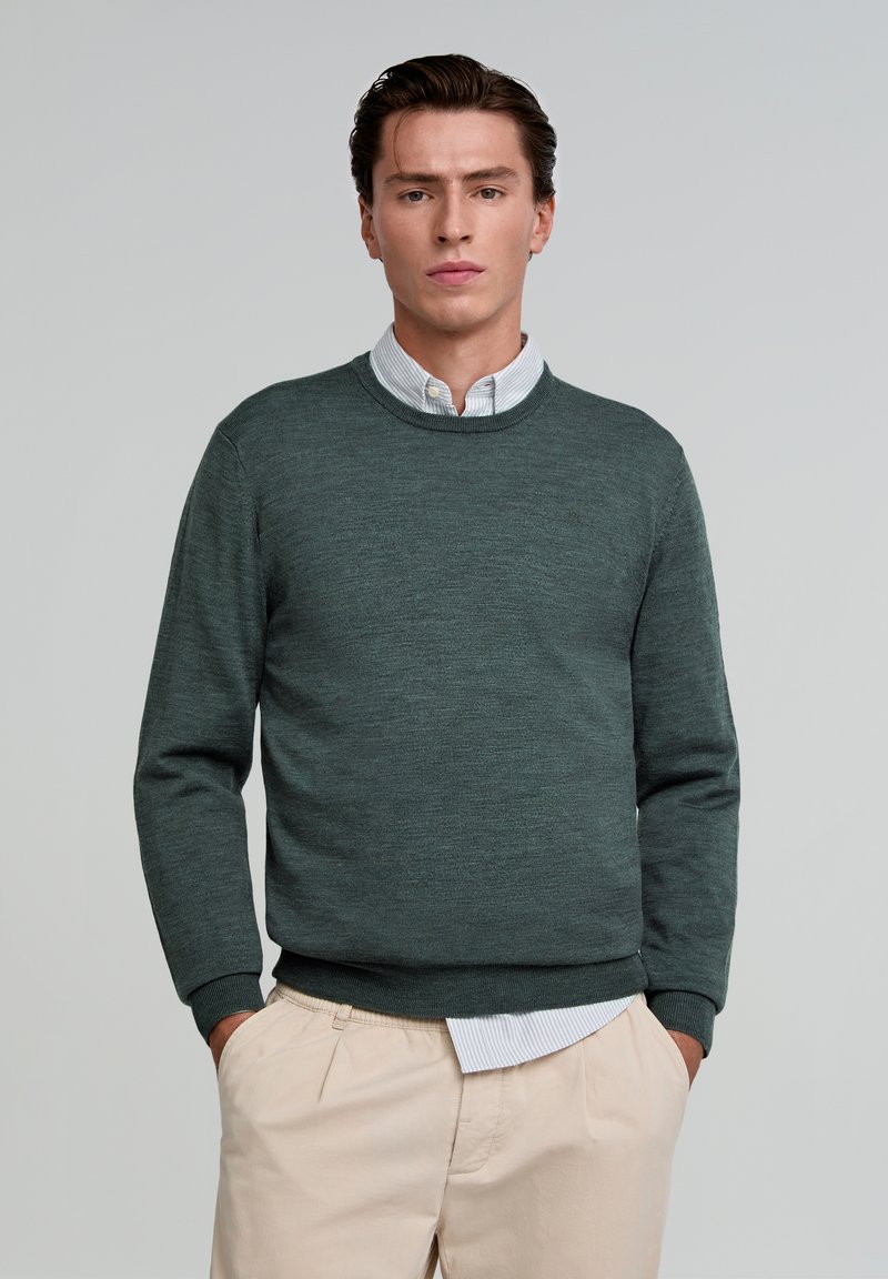 Donkergroen crewneck trui gemaakt van gestructureerde stof, met lange mouwen en een ribbelrand, gedragen over een lichtblauw overhemd met kraag.