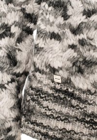 Maglione lavorato a maglia con un motivo grigio e bianco, caratterizzato da una consistenza spessa. Include un piccolo tag metallico con logo all'orlo.
