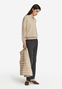 Beige V-Ausschnitt Pullover aus weichem Material, kombiniert mit dunkelgrauen Jeans. Hält eine leichte, gesteppte ärmellose Jacke. Braune Blockabsatz-Mules.