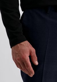 Mannelijke hand die rust naast donkerblauwe geruite broek en een zwart overhemd met lange mouwen, en de natuurlijke huidtextuur en ontspannen vingers toont.