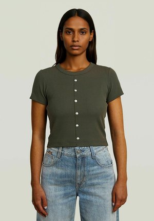 Femme aux longs cheveux foncés portant un haut boutonné vert olive à manches courtes et un jean taille haute bleu clair sur un fond uni.