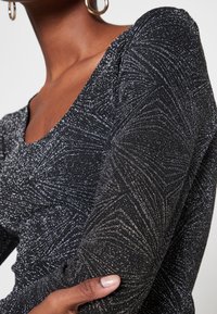Frau in einem schwarzen, glitzernden Langarmtop mit einem strukturierten, funkensprühenden Wirbelmuster und goldenen Creolen, eine Hand ruht auf dem Arm.