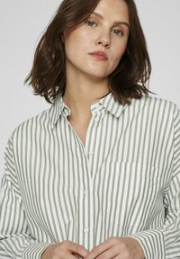 Camisa de botón hecha de algodón suave, con rayas verticales verdes y blancas, un bolsillo en el pecho y un cuello puntiagudo.