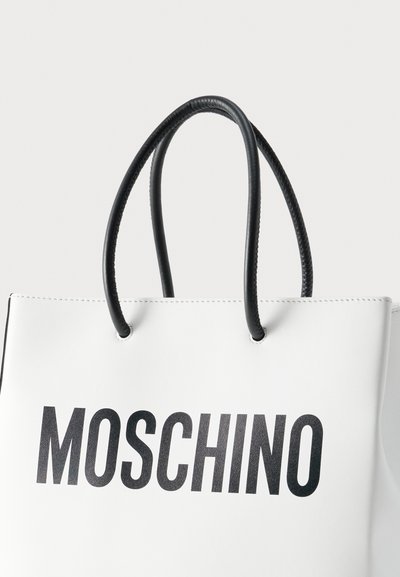 Λευκή τσάντα ώμου με μαύρες στρογγυλεμένες λαβές και έντονο μαύρο λογότυπο "MOSCHINO" μπροστά πάνω σε μονόχρωμο φόντο.