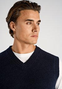 Marineblauer Strickweste mit V-Ausschnitt und geripptem Rand, getragen über einem weißen Hemd. Strukturierter Stoff, figurbetontes Design, minimalistische Details.