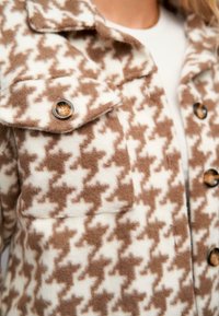 Jachetă din polar cu model houndstooth maro și alb, prevăzută cu un buzunar cu clapetă și nasturi întunecați. Material texturat cu un aspect moale.