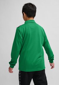 Groene langekousen polo met een textuurstof, met een kraag en zwarte accenten op de schouders. Draagt een zwarte broek.