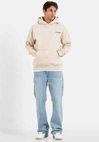 Sudadera beige con un bolsillo frontal y un logo verde, combinada con jeans azul claro y zapatillas blancas, con un corte relajado y una textura suave.