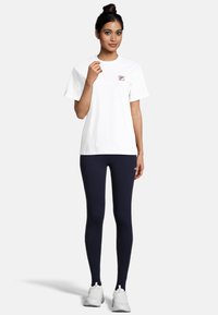 Witte katoenen t-shirt met een klein logo op de linkerborst, gecombineerd met marineblauwe legging. Model draagt witte sportschoenen en staat in een neutrale pose.