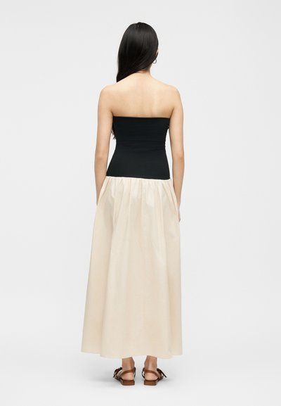 Γυναίκα με μακριά σκούρα μαλλιά, φαινομένη από πίσω, φοράει ένα μαύρο στράπλες τοπ, εκρού φουσκωτή φούστα και καφέ σανδάλια με επίπεδη σόλα.