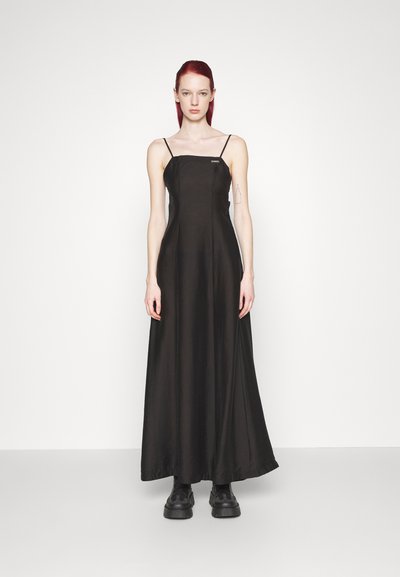 Čierne maxi šaty s tenkými ramienkami, hladkým povrchom a priliehavým živôtikom. Nosí sa s čiernymi platformovými topánkami, vystavené na neutrálnom pozadí.