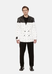 Dwurzędowa marynarka w białym kolorze z czarnymi skórzanymi akcentami na ramionach i kołnierzu, zdobiona powtarzającym się nadrukiem małych symboli. Czarne spodnie dopełniają całość.