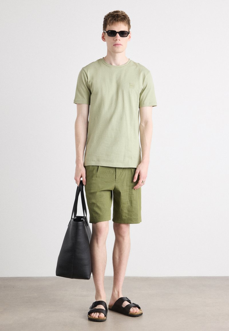 T-shirt en coton vert olive clair et shorts assortis en olive. Le modèle tient un sac en cuir noir, porte des lunettes de soleil noires et des sandales.
