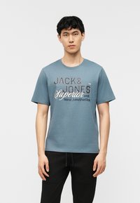 Jeune homme debout, portant un t-shirt bleu de la marque Jack & Jones et un pantalon noir, sur un fond blanc uni.