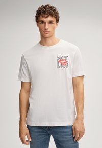 Jeune homme aux cheveux bouclés portant un t-shirt blanc avec le texte "SOHO LOVE" et un graphique de lèvres rouges sur la poitrine, associé à un jean bleu.