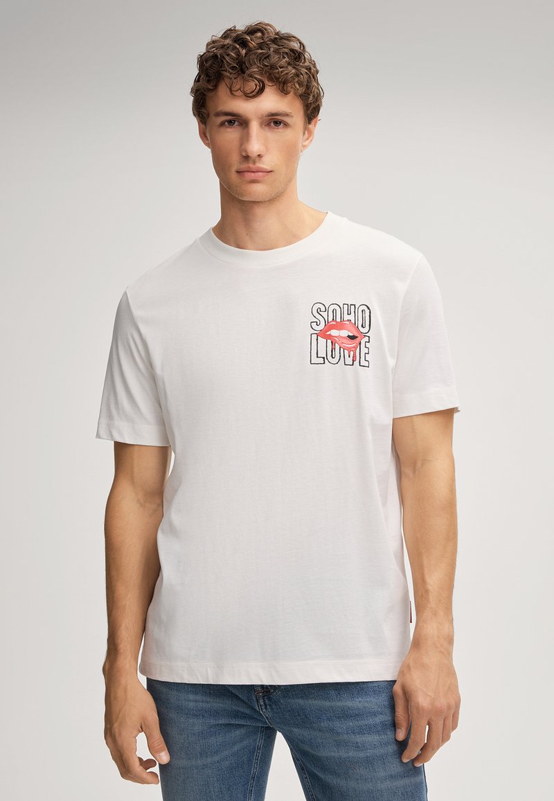Jeune homme aux cheveux bouclés portant un t-shirt blanc avec le texte "SOHO LOVE" et un graphique de lèvres rouges sur la poitrine, associé à un jean bleu.