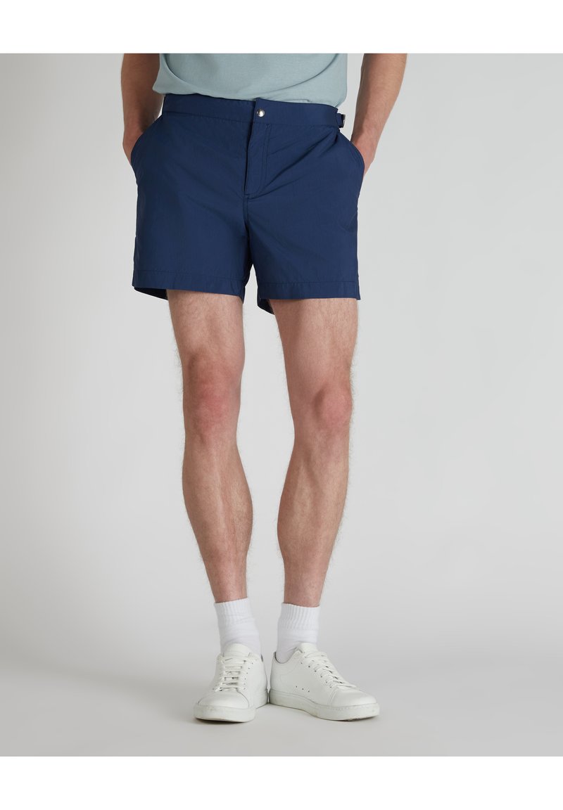 Marineblaue Shorts aus leichtem Material mit flachem Bund, seitlichen Taschen und Knopfverschluss. Tragen kombiniert mit weißen Turnschuhen und Socken.