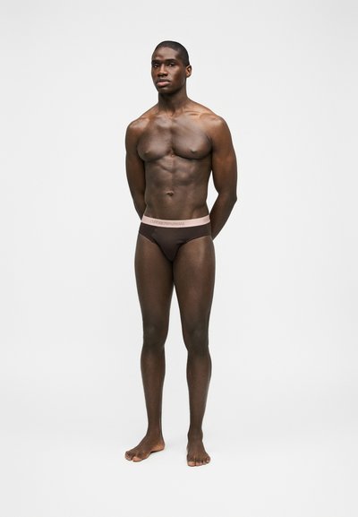 Μυώδης άνδρας που στέκεται ξυπόλητος, φορώντας καφέ εσώρουχο με ανοιχτό ροζ ζώνη μέσης Emporio Armani, σε απλό λευκό φόντο.