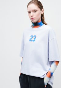 Svetlošedé oversized tričko s modrým a oranžovým grafickým dizajnom s nápisom "Jordan 23." Model má na sebe farebnú blúzku s vysokým golierom.