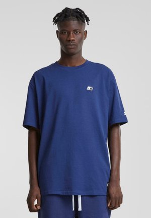 T-shirt en coton bleu marine avec un col rond, des manches courtes et un petit patch logo sur la poitrine. Design minimaliste et coupe décontractée.
