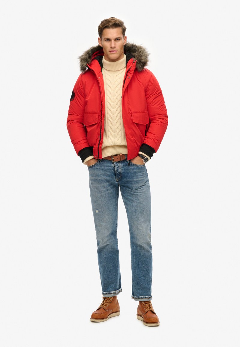 Doudoune rouge avec capuche en fourrure, deux poches avant, portée par-dessus un pull en tricot beige à mailles, associée à un jean bleu et des bottes marron.