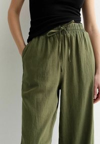 Olive green, loose-fitting pants made of textured fabric, featuring an elastic waistband with a drawstring and side pockets.