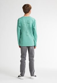 Langärmliges, mintgrünes Baumwoll-T-Shirt mit einem Baum-Graphic und "PETROL"-Text auf dem Rücken, getragen mit grauen Hosen und weißen Sneakers.
