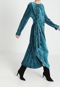 Tyrkysové sametové šaty s vertikálním vroubkovaným vzorem, dlouhými rukávy a zavázaným pasem. Vysoký rozparek a kombinované s černými špičatými kotníkovými botami.
