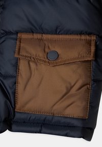 Sección de chaqueta acolchada con un exterior azul marino y un bolsillo cuadrado marrón. El bolsillo cuenta con un cierre de botón con un logo de la marca.
