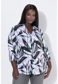 Žena s dlouhými rovnými vlasy, oblečená v bílé blůze se vzory zelených a fialových listů a šedých kalhotách, stojící na bílém pozadí.