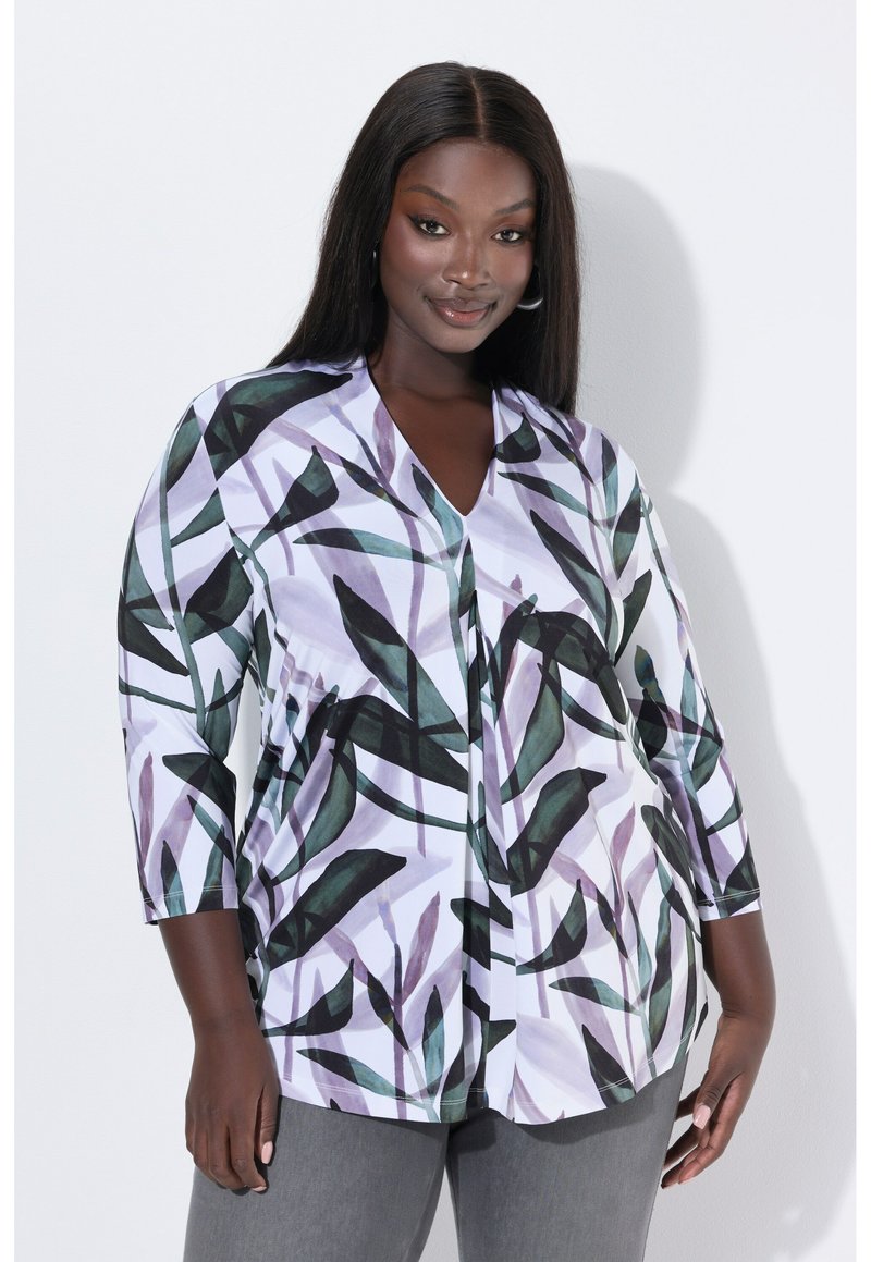 Žena s dlouhými rovnými vlasy, oblečená v bílé blůze se vzory zelených a fialových listů a šedých kalhotách, stojící na bílém pozadí.