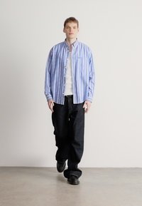 Svītraina zilā un baltā pogājama krekls pār baltu džemperi, kombinēts ar vaļīgām melnām džinsām un melniem apaviem. Vienkāršs fons.