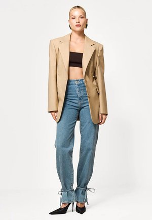 Kobieta nosząca beżową, oversize marynarkę, czarny krótki top, wysokie niebieskie jeansy z wiązaniami przy kostkach oraz czarne szpiczaste szpilki, stojąca prosto.