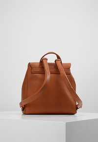 Brun læderrygsæk med glat tekstur, struktureret form, dobbelte stropper og et top håndtag. Har minimal syning og et strømlinet design.