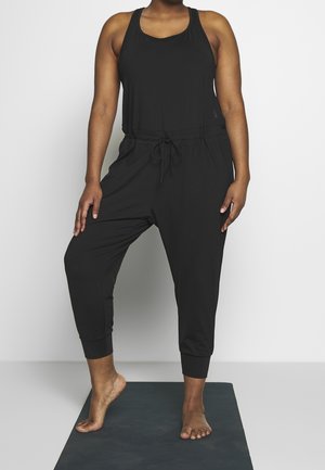 Persoon die een zwarte mouwloze jumpsuit draagt, staat op één been op een donkere fitnessmat tegen een effen achtergrond, op blote voeten.