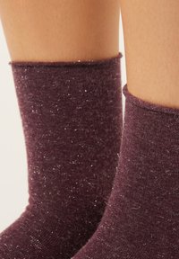 Bordó ponožky z měkké, třpytivé látky. Mají žebrovaný manžet a jemný lesk po celé ploše, s těsným přilnutím kolem kotníku.