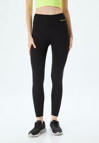 Zwarte legging met een hoge tailleband, gemaakt van rekbaar materiaal, met een klein, geel "REALITY" tekstdetail op de tailleband.