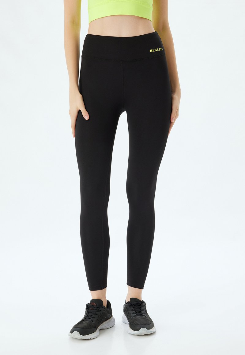 Zwarte legging met een hoge tailleband, gemaakt van rekbaar materiaal, met een klein, geel "REALITY" tekstdetail op de tailleband.