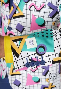 Giacca elegante colorata con un motivo a griglia e forme geometriche in rosa, verde, viola e giallo. Bottoni blu e etichetta di marca visibili.