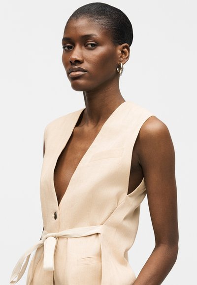 Femme à la peau foncée et aux cheveux courts portant un gilet beige sans manches avec un col en V profond et une ceinture nouée, sur un fond clair uni.