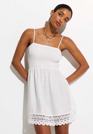 Vrouw draagt een mouwloze witte jurk met geplooid lijfje en kanten zoom, poseert met handen achter de rug tegen een witte achtergrond.