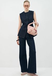 Denim vest met een hoge halslijn en een sluiting met knopen aan de voorkant, gecombineerd met een ruime donkerdenim broek. Lichtroze handtas van geweven materiaal en zonnebril.