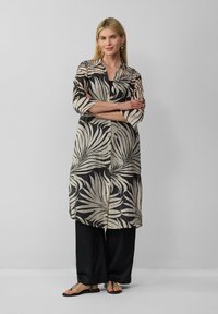 Blusen-Tunika mit schwarzem und beigem Blattmuster, langen Ärmeln und seitlichen Schlitzen, kombiniert mit schwarzen weit geschnittenen Hosen und Sandalen.