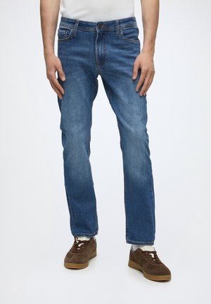 Homme portant un jean bleu, des chaussettes blanches et des baskets marron, debout devant un fond clair uni.