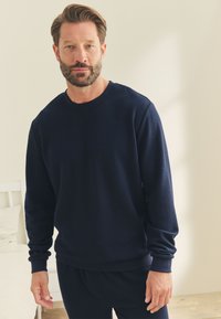 Mand med kort brunt hår og skæg iført en mørkeblå, langærmet, tekstureret sweater, der står indendørs nær en hvid seng.