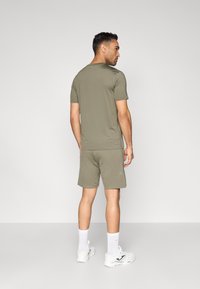 Ανδρικό αθλητικό σύνολο: κοντομάνικη μπλούζα σε ελιά πράσινο και αντίστοιχα σορτς, κατασκευασμένα από απαλό ύφασμα· συνδυασμένο με λευκά αθλητικά παπούτσια και κάλτσες.