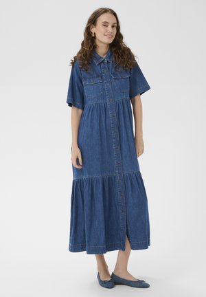 Kobieta ubrana w długą, niebieską sukienkę dżinsową na guziki z krótkimi rękawami i kołnierzykiem, w parze z niebieskimi balerinami, stojąca na jednolitym tle.