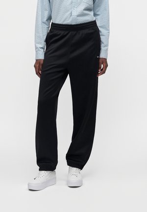 Person iført sorte løstsiddende bukser, lyseblå mønstret langærmet skjorte og hvide platformsneakers, stående foran en ensfarvet baggrund.