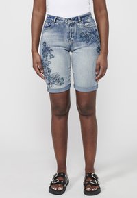 Shorts en denim bleu clair avec broderies florales en bleu foncé et vert, dotés de ourlets retroussés et d'une coupe slim. Sandales noires avec des accents.
